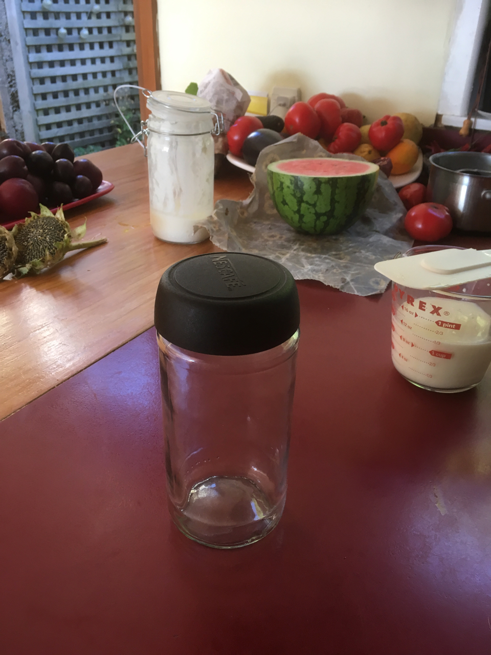 An clean unlabelled glass jar with a plastic lid sitting on a kitchen bench. There are various fruits in bowls behind it. Directly behind the empty jar there is a glass jar which contains yoghurt. It is nearly empty. To the right there is a 500ml jug which is half full of milk.